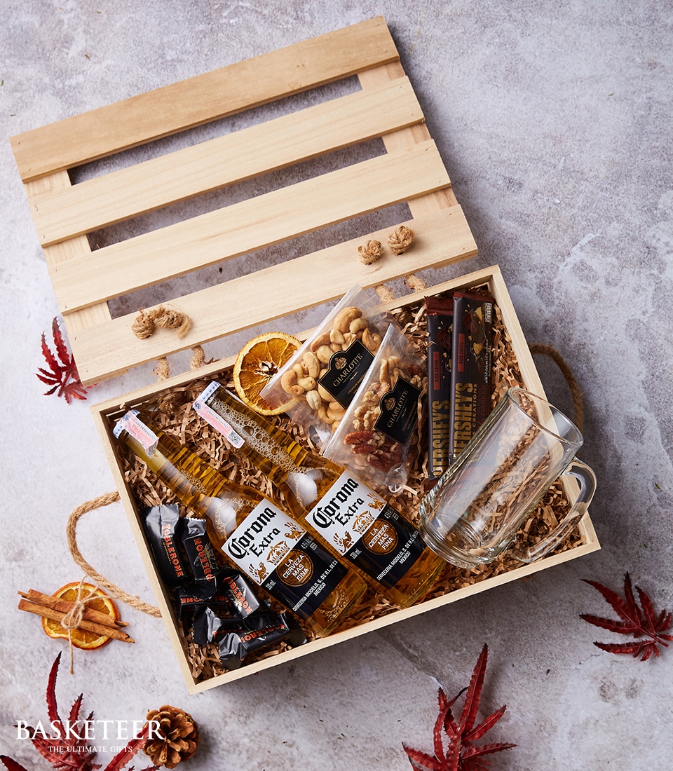 A luxurious wooden crate gift box from Basketeer, adorned with their signature sliding lid, presents a curated array of indulgent delights. Nestled within the box, you'll find elegantly packaged Corona beer bottles alongside a crystal-clear beer mug that promises to elevate your drinking experience. Complementing these are gourmet packets of mixed nuts and artisanal snack sticks, all cushioned in rich wooden wool packing material. This sophisticated scene is set against a light textured backdrop, accented by scattered autumn leaves that evoke a sense of warmth and seasonal charm. The Basketeer logo graces the ribbon and box, marking this offering as one of refined taste and elegance.