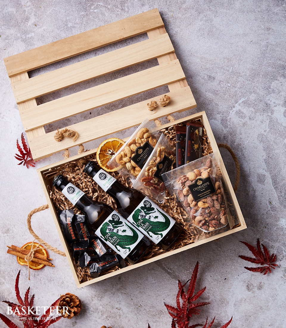 A beautifully crafted wooden gift box from Basketeer with its lid gracefully open, unveiling a lavish assortment of gourmet delights. Nestled within the straw filling are two premium beer bottles, artisan packets of nuts, hand-picked dried fruits, and succulent sausage sticks. Surrounding this luxurious spread are autumn leaves, adding a touch of seasonal elegance to the scene. The box proudly displays the distinguished 