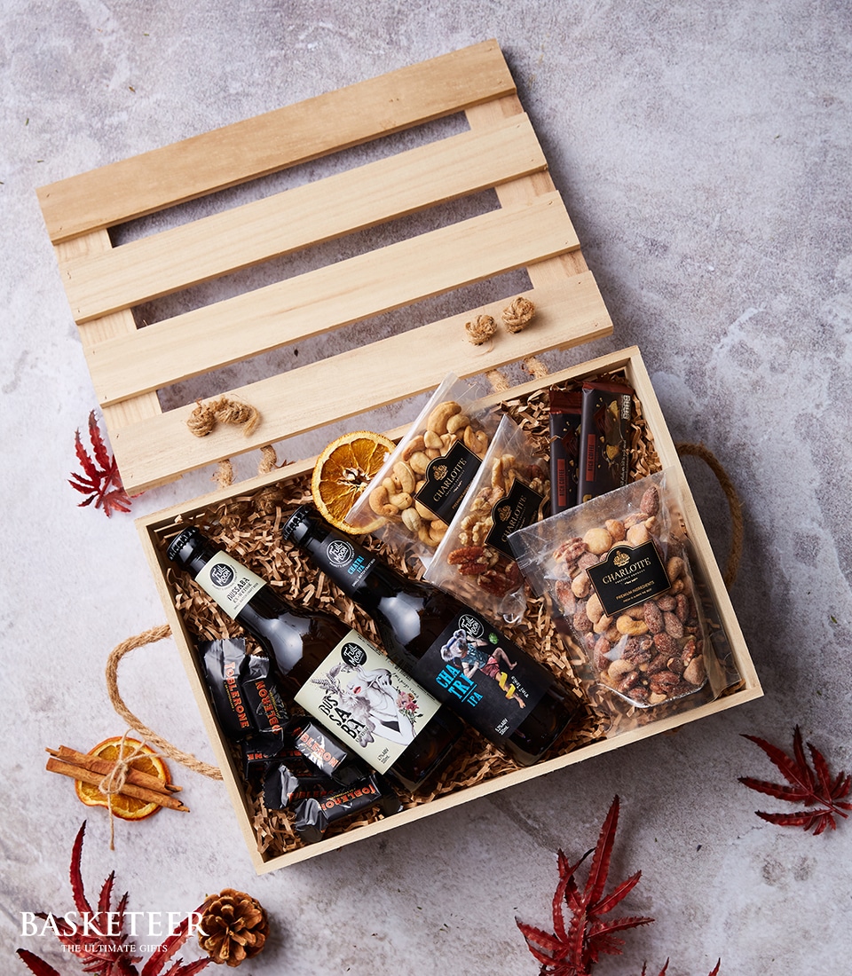 An elegant wooden gift box lies open, inviting you to indulge in its luxurious contents. Inside, you'll find a curated selection of premium craft beers, an assortment of gourmet mixed nuts, decadent chocolates, and a delightful bag of artisan popcorn. The box is nestled in soft shredded paper and beautifully adorned with dried orange slices and autumn leaves, creating a cozy yet sophisticated scene. A tasteful ribbon graced with the Basketeer logo adds the final touch to this exquisite presentation.