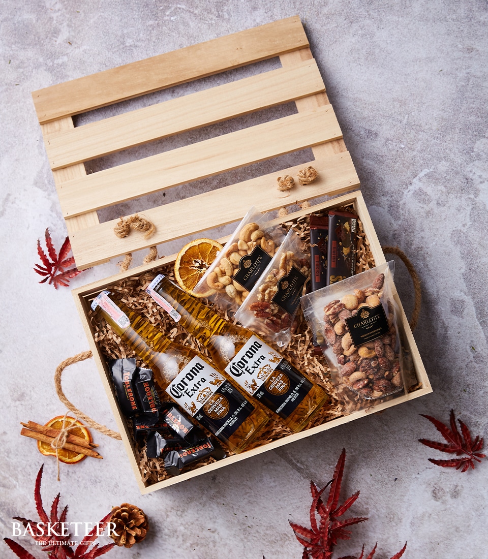 A lavish wooden crate brimming with four bottles of Corona Extra beer, nestled amidst gourmet packets of nuts and delectable sausage sticks, all artfully arranged on a bed of rustic straw. The lid lies elegantly behind the open box, revealing every detail. Embellished with decorative autumn leaves and a sophisticated rope handle, this scene celebrates indulgence and luxury. Text on the side proudly declares 