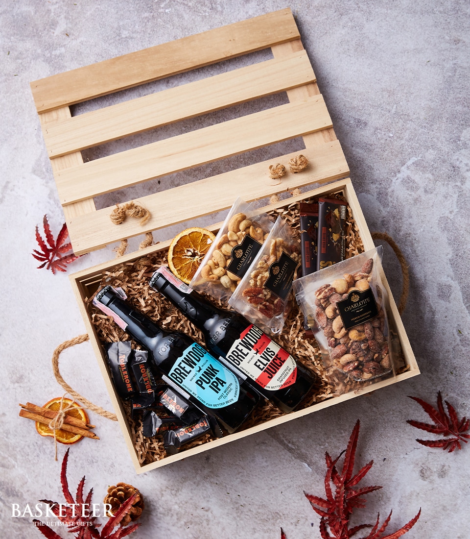 A luxurious wooden gift box from Basketeer, elegantly open to reveal a curated selection of gourmet indulgences. Inside, discover two premium bottles of Brewdog beer nestled alongside artisanal packs of nuts, decadent chocolate bars, and exquisite dried citrus slices. The Basketeer-branded ribbon and packaging add a touch of sophistication to this sumptuous assortment. The box is artfully displayed on a pristine marble surface adorned with vibrant red maple leaves, enhancing the inviting ambiance of this extraordinary gift.