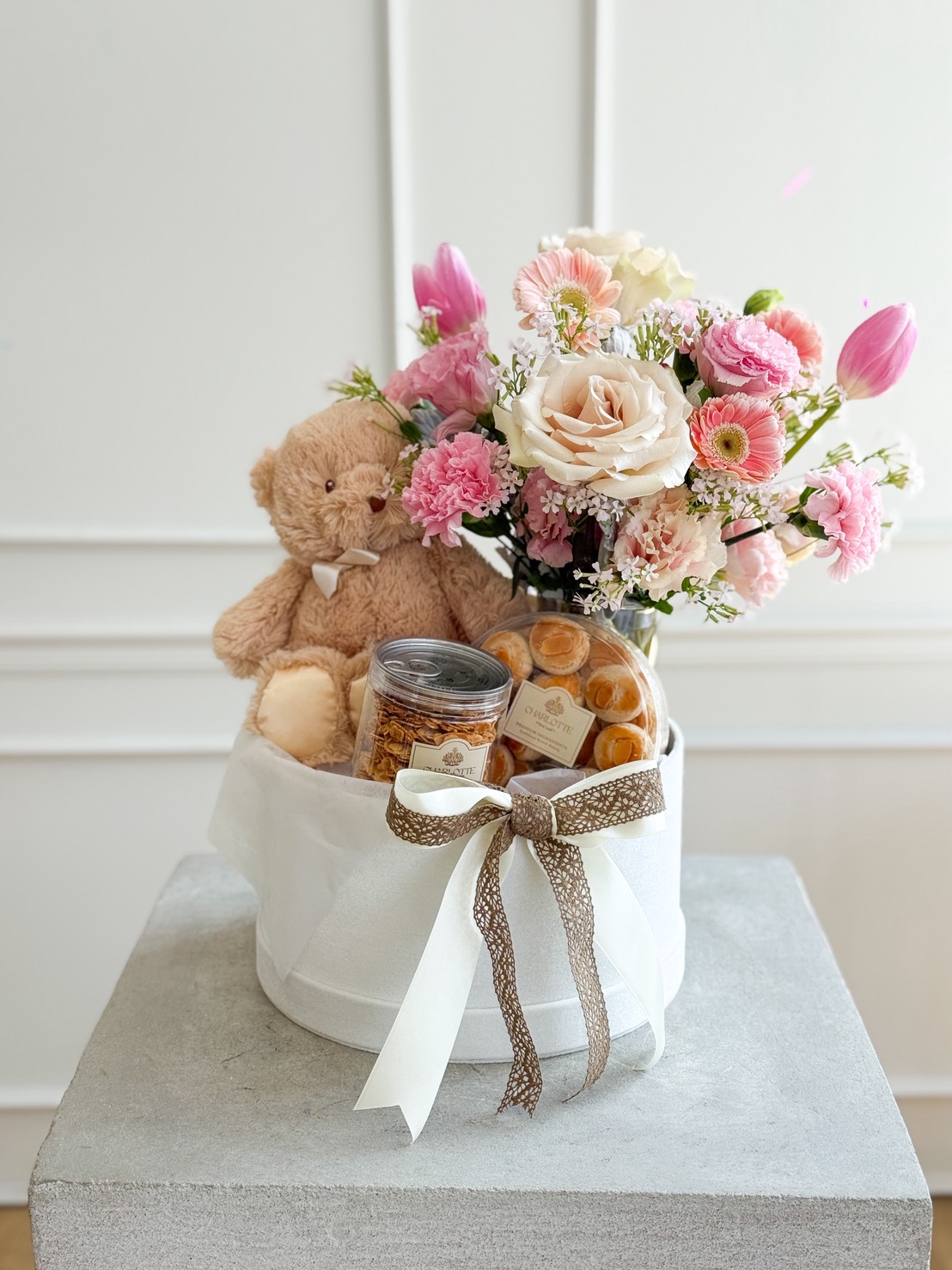 A white gift box with a brown and white ribbon, containing a plush teddy bear, jars of snacks, and a bouquet of pink and white flowers, displayed on a grey pedestal against a white wall.