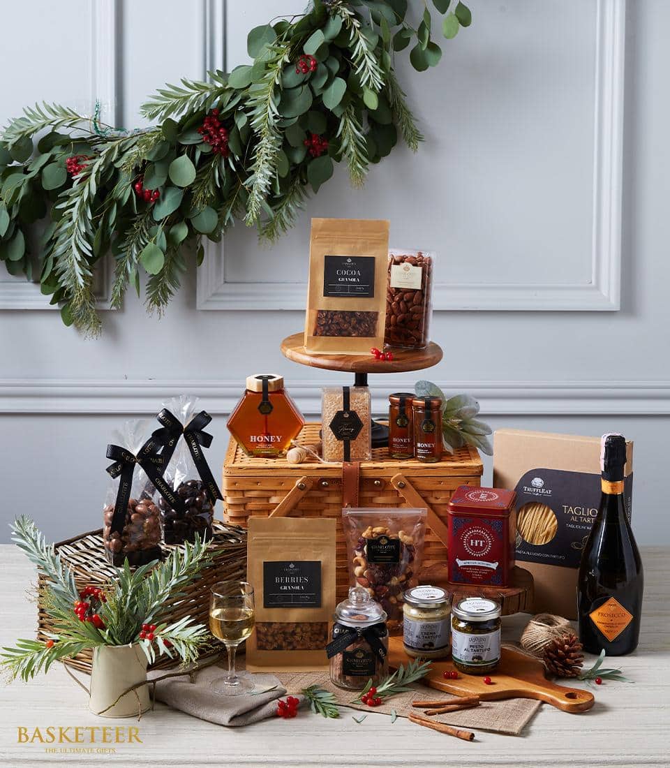 A lavish holiday gift basket by Basketeer, brimming with an exquisite selection of gourmet delights—luscious honey, premium nuts, decadent chocolates, rich sauces, and a fine bottle of wine. The basket is elegantly garnished with lush greenery and vibrant red berries. Against a pristine white wall adorned with a stunning garland above, the scene exudes sophistication and festive charm. The signature Basketeer ribbon artfully completes this luxurious presentation.