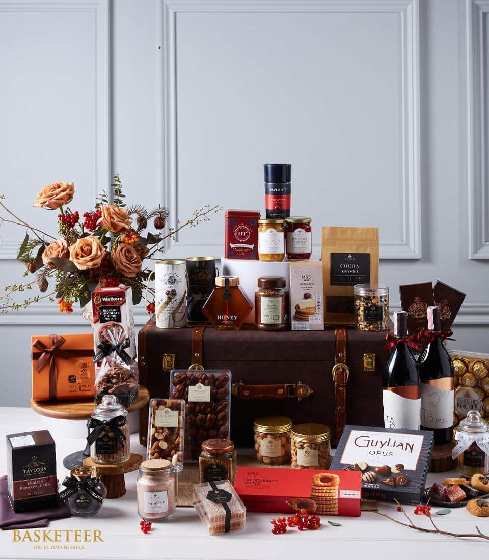 A sumptuous Basketeer gift hamper luxuriously displayed atop and around an exquisite vintage suitcase. This opulent assortment features premium wine bottles, artisanal honey, fine teas, decadent chocolates, exquisite jam, gourmet nuts, and an array of other delectable treats. A vase of elegantly arranged dried flowers graces the left side, enhancing the sophisticated ambiance. Ribbons and boxes adorned with the prestigious Basketeer logo add a touch of distinguished charm to this inviting scene.