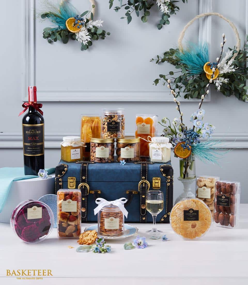 A sumptuous Basketeer gift hamper exuding luxury and opulence. The ensemble includes an exquisite bottle of red wine, gourmet jars of premium nuts, artisanal biscuits, delectable dried fruits, and indulgent confectionery. Each item is meticulously arranged on and around a sophisticated blue trunk adorned with a chic ribbon bearing the Basketeer logo. The backdrop showcases elegant floral decorations against a tasteful grey wall, amplifying the sense of refinement and exclusivity.