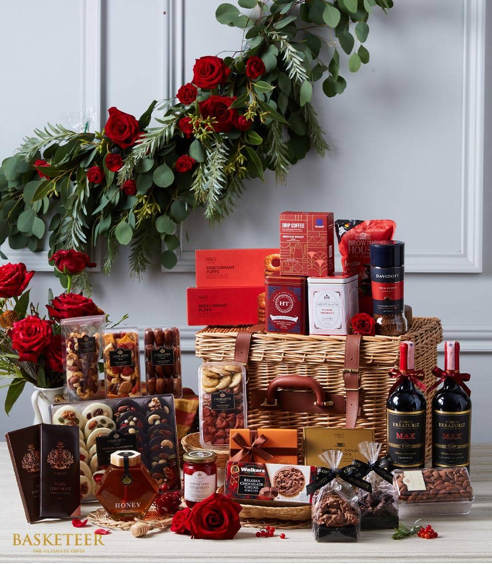 An opulent Basketeer gift basket, elegantly adorned with lush red roses and vibrant greenery, exudes luxury against a pristine white wall. The meticulously arranged assortment of gourmet delights includes two exquisite wine bottles, decadent chocolates, artisanal cookies, premium nuts, rich coffee, and golden honey. Each item is thoughtfully placed within and around the sophisticated wicker basket. A Basketeer-branded ribbon gracefully encircles the arrangement, highlighting the brand's commitment to elegance and refinement.