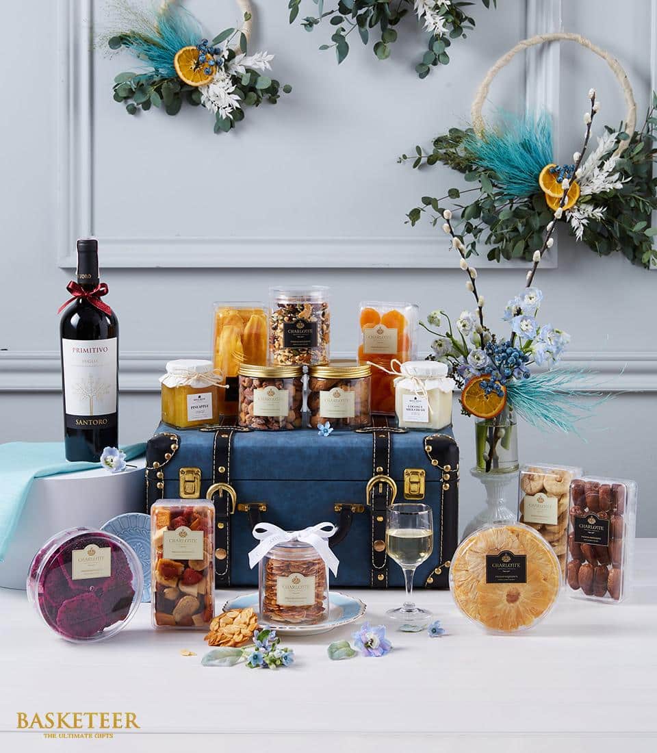Luxurious gift set by Basketeer showcased on a sophisticated blue chest, featuring an exquisite bottle of wine, artisanal jars of jam, and a selection of gourmet dried fruits and nuts. The display is beautifully embellished with delicate flowers and lush greenery, all accented in gold for an opulent touch. Elegant packaging proudly displays the Basketeer logo on ribbons and boxes, creating an inviting scene against a chic light gray backdrop.