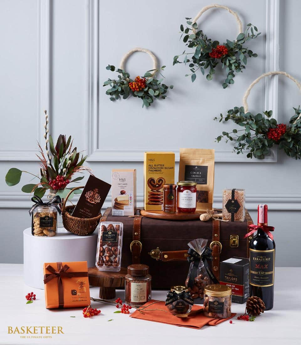 A luxurious gift hamper is exquisitely arranged in front of a sophisticated gray paneled wall adorned with three elegant festive wreaths. The Basketeer-branded ribbon graces the items within, amplifying the sense of opulence. This indulgent collection features a bottle of fine red wine, artisanal chocolates, premium nuts, delectable jams, and gourmet biscuits, all meticulously presented around a chic leather suitcase. Perfect for those who appreciate the finer things in life.