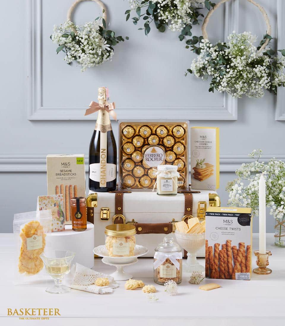 A luxurious gift hamper branded with the Basketeer logo elegantly displayed atop a refined white and brown suitcase. This indulgent assortment includes artisan crackers, savory cheese twists, a jar of golden honey, and an inviting scented candle. Decadent Ferrero Rocher chocolates, a bottle of exquisite champagne, and a crystal glass filled with premium white wine complete this sumptuous offering. The background is beautifully adorned with wreaths and flowers, enhancing the opulent ambiance of this sophisticated scene.