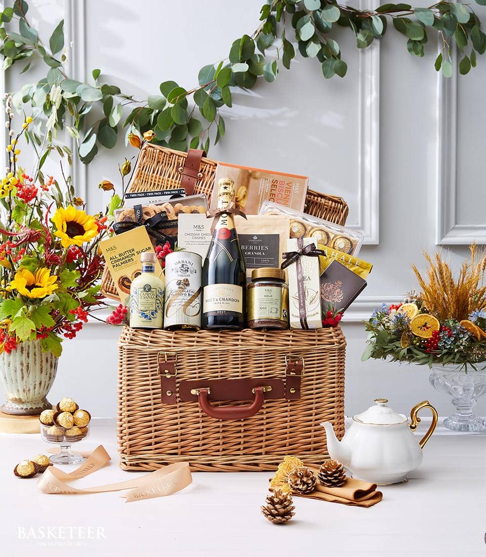 An opulent wicker basket from Basketeer, brimming with an exquisite selection of gourmet treasures including a fine bottle of wine, decadent chocolates, buttery biscuits, and artisanal preserves, is elegantly showcased on a lavish table. The scene is adorned with lush floral arrangements and a pristine white teapot, complemented by a luxurious ribbon emblazoned with 