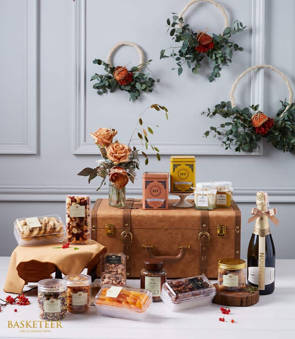 A lavish and inviting display featuring a luxurious leather trunk from Basketeer, exquisitely filled with an assortment of gourmet indulgences. Delight in an array of fine cheeses, premium nuts, artisanal cookies, delectable jam and honey, all complemented by an elegant bottle of sparkling wine. Fresh flowers and festive wreaths adorned with dried orange slices enhance the ambiance. The BASKETEER logo elegantly graces the bottom left corner, signifying exceptional quality and sophistication.