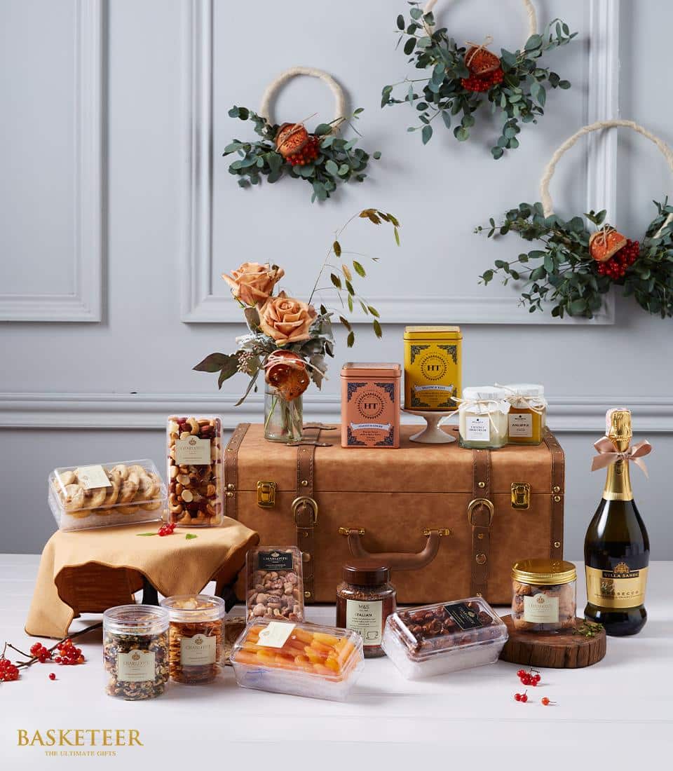 A luxurious display showcases a refined leather suitcase, beautifully arranged with an assortment of gourmet snacks and beverages. The selection includes fine nuts, exquisite dried fruits, rich spreads, and a distinguished bottle of wine. A sophisticated touch is added by a vase filled with vibrant orange roses and lush eucalyptus leaves. Elegantly draped wreaths with red accents adorn the sophisticated gray wall in the background. The Basketeer brand's logo on an enchanting ribbon elegantly tied around the suitcase elevates the entire scene into an inviting celebration of taste and style.