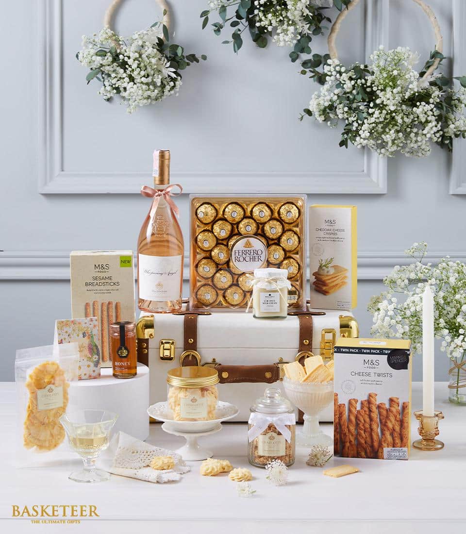A lavish gift arrangement by Basketeer is presented, showcasing a premium bottle of rosé, delectable Ferrero Rocher chocolates, gourmet M&S snacks, and a jar of artisanal honey. Each item is thoughtfully arranged on and around an exquisite white suitcase adorned with gold accents. In the background, elegant floral decorations enhance the scene's sophistication. A ribbon featuring the Basketeer logo adds an inviting touch of elegance to this luxurious display.