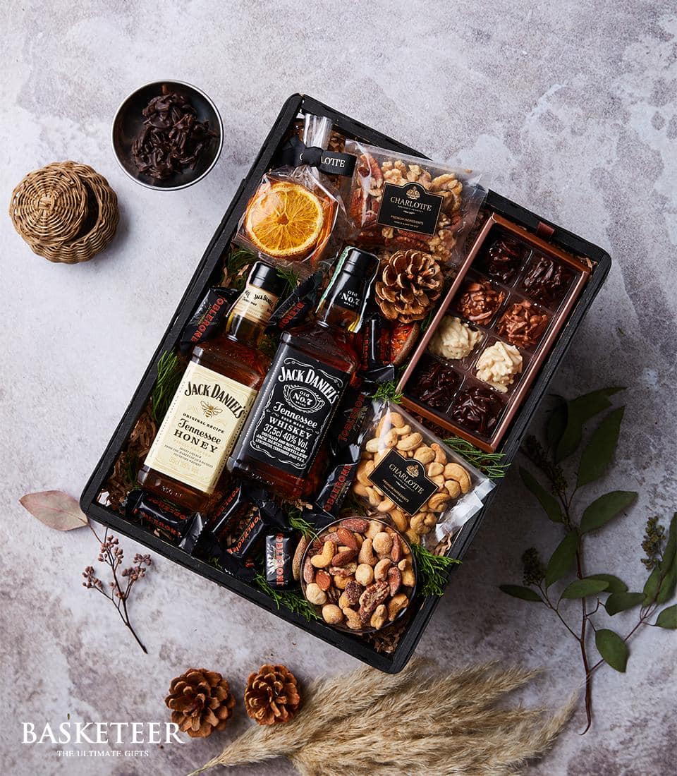 A luxurious Basketeer gift basket is nestled on a sophisticated gray textured surface, adorned with the distinguished Basketeer logo. Inside, two exquisite bottles of Jack Daniel's whiskey take center stage, surrounded by an opulent assortment of gourmet delights. Indulge in premium nuts, rich chocolate pieces, and decadent assorted truffles that promise to tantalize your taste buds. Adding an elegant touch are dried orange slices displayed in a charming bowl alongside decorative pinecones. This meticulously curated collection invites indulgence and celebrates the art of gifting with unmatched sophistication.
