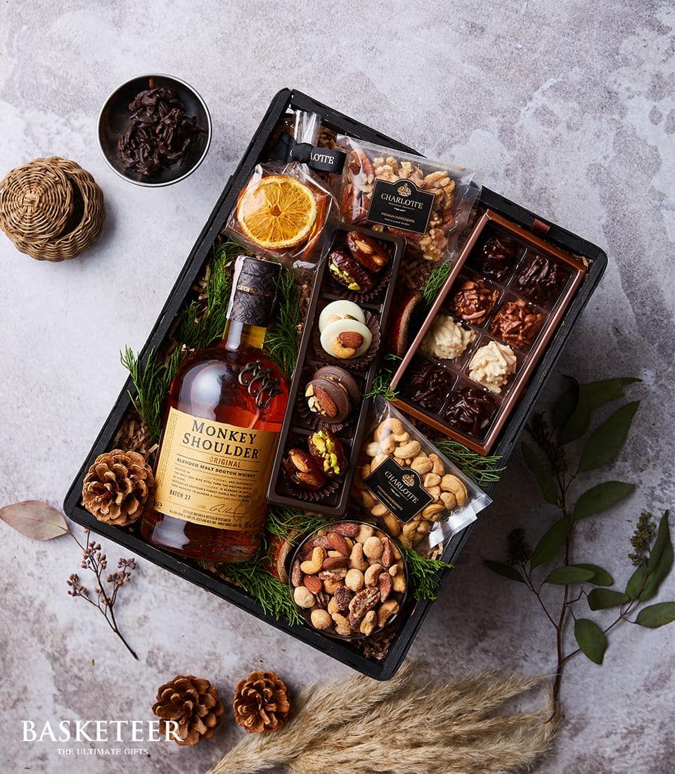 A top-down view of an exquisite gift basket curated by Basketeer, featuring a premium bottle of Monkey Shoulder whisky surrounded by a lavish assortment of gourmet nuts, dried fruits, and decadent chocolates. The basket is elegantly adorned with pine cones and lush fir branches, creating a harmonious blend of rustic charm and sophisticated luxury. Set against a textured gray surface, the scene is complete with the distinguished 