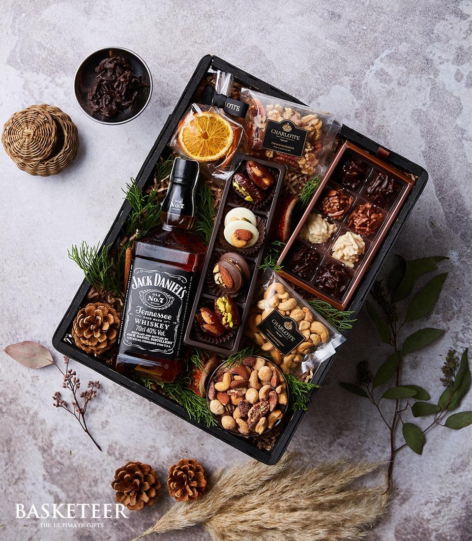 Basketeer gift basket showcasing a premium bottle of Jack Daniel's Tennessee Whiskey, elegantly nestled among an array of gourmet delights: artfully dried orange slices, a selection of fine nuts, rich chocolates, and luscious dried fruits. Each delicacy is thoughtfully placed within its own compartment. The scene is beautifully adorned with rustic pine cones and delicate sprigs of dried wheat for a sophisticated touch. Everywhere you look, the Basketeer logo promises quality and luxury.