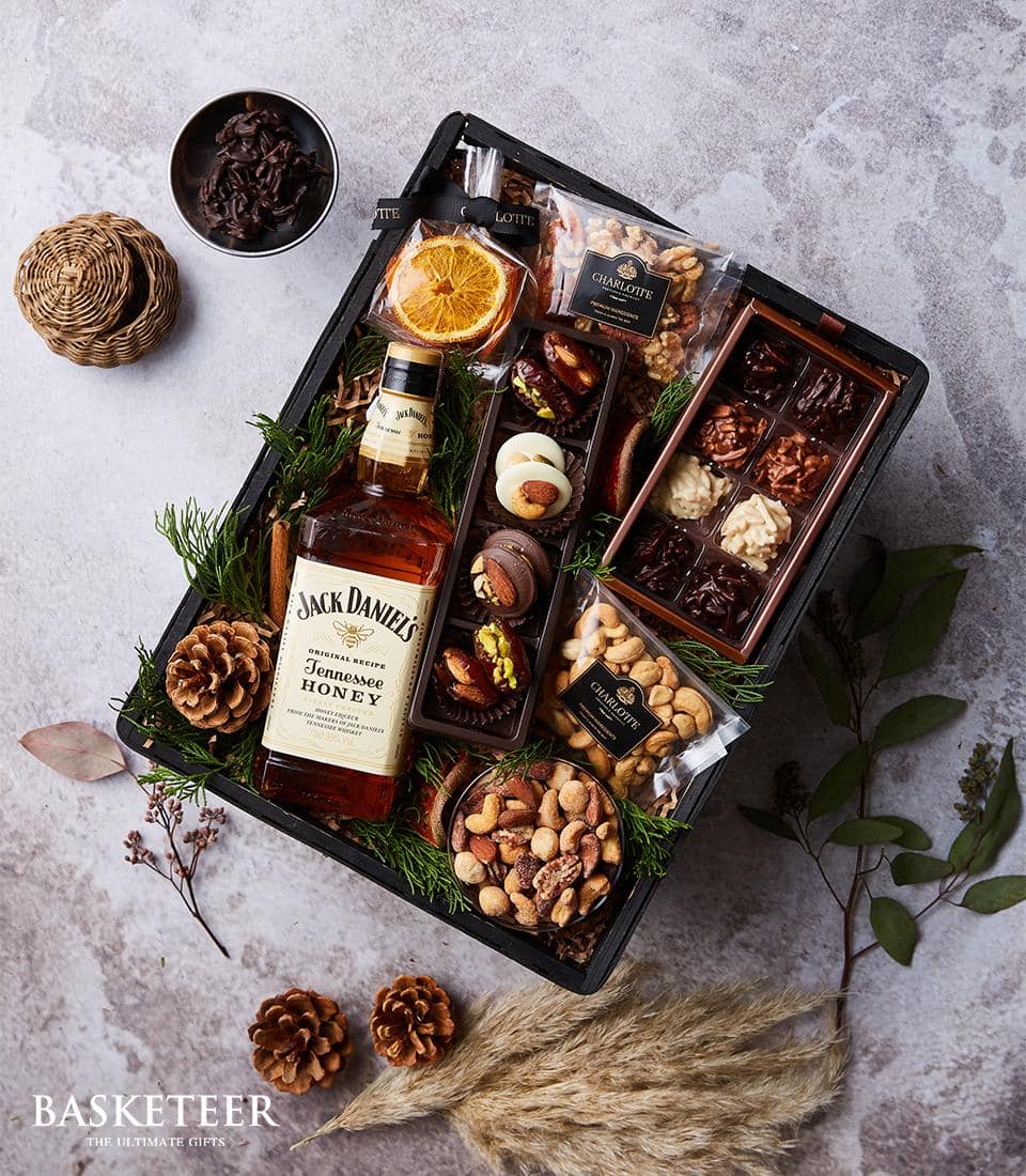 A luxurious gift basket curated by Basketeer, featuring a bottle of Jack Daniel's Tennessee Honey whiskey, premium assorted chocolates, gourmet nuts, exquisite dried fruits, a charming jar of artisanal honey, and decadent macaroons. The basket is elegantly adorned with festive pinecones, lush green foliage, and aromatic dried orange slices. Delicately tied with a sophisticated ribbon showcasing the Basketeer logo.