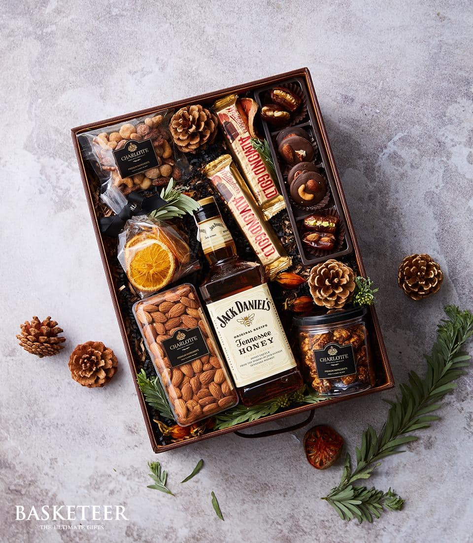 A lavish gift basket brimming with an exquisite selection of treats awaits. Nestled within, you’ll discover a bottle of Jack Daniel's Tennessee Honey, its golden label gleaming invitingly. Surrounding it are decadent chocolate-covered almonds and a medley of whole almonds. Dried orange slices add a touch of rustic elegance, while a premium bag of assorted nuts offers savory delight. A petite honey jar glistens with natural sweetness, complemented by an array of luxurious chocolate snacks. The ensemble is beautifully adorned with ribbons and the distinguished Basketeer logo in the bottom left, ensuring this gift embodies both indulgence and sophistication.