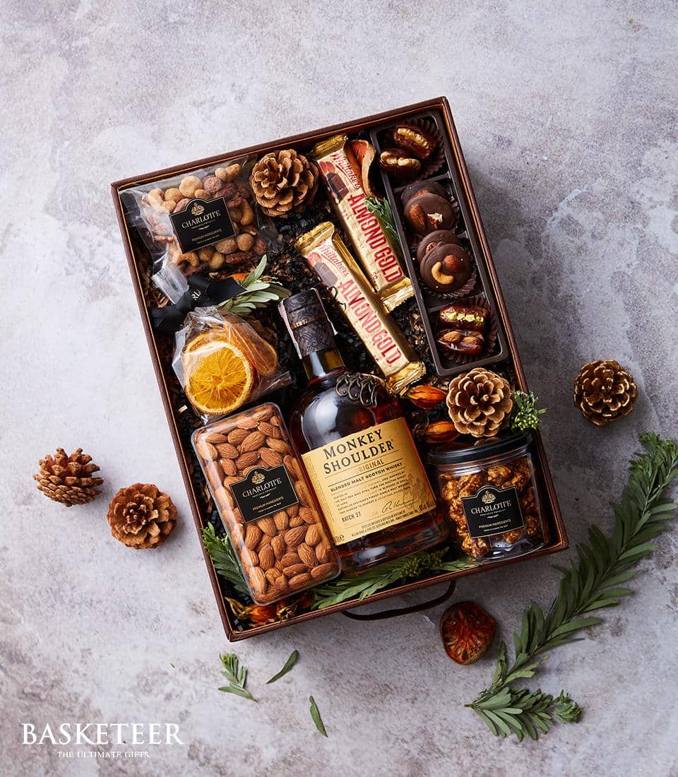 A luxurious gift box from Basketeer featuring a bottle of premium Monkey Shoulder whisky, elegantly accompanied by gourmet jars of snacks and an exquisite selection of assorted chocolates. Adorned with a dried orange slice and festively arranged pine cones, the compartmentalized display exudes sophistication with green foliage accents. The Basketeer logo graces the ribbon, adding a touch of brand distinction to this inviting presentation.
