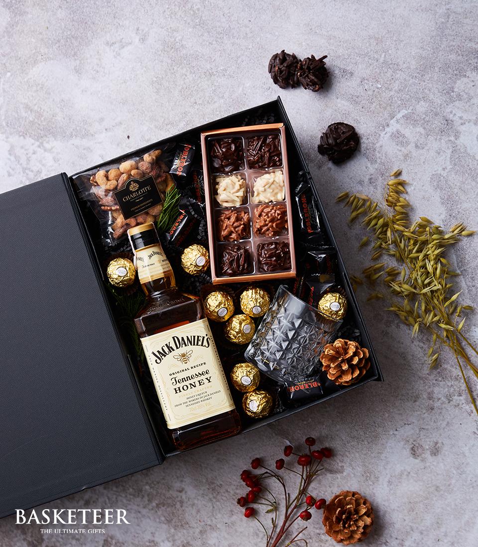 A luxurious Chocolates & Whiskey Gift Set from Basketeer, elegantly showcasing a bottle of Jack Daniel's Tennessee Honey whiskey paired with an exquisite assortment of chocolates and Ferrero Rocher delights. The set is thoughtfully adorned with delicately candied nuts, charming small decorations, and lush sprigs of greenery intertwined with vibrant red berries. Presented on a sophisticated textured surface, the gift set exudes refinement and opulence. A ribbon bearing the Basketeer logo is gracefully tied around the elegantly designed box, completing this lavish presentation.