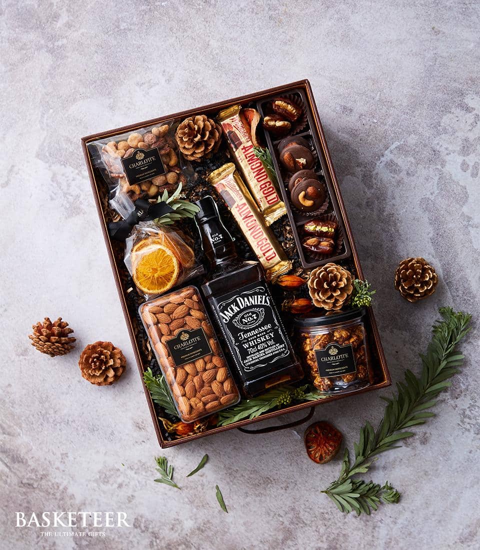 A lavish holiday gift basket from Basketeer, showcasing a distinguished bottle of Jack Daniel's whiskey nestled among an array of gourmet chocolates, premium nuts, and elegantly dried orange slices. This exquisite selection is artistically adorned with festive pine cones and lush greenery. The luxurious arrangement rests on a textured gray surface, with the prestigious BASKETEER logo elegantly displayed in the bottom left corner.