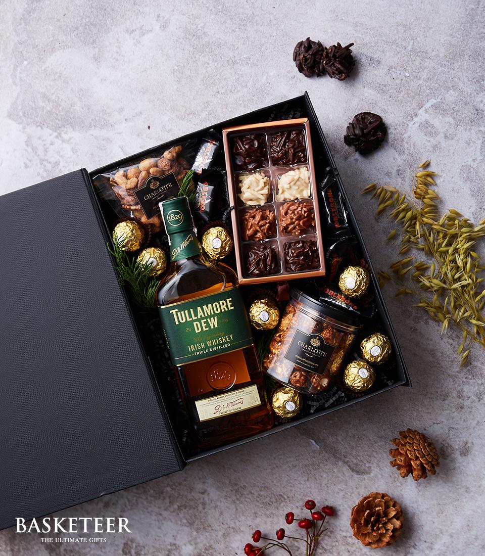 A lavish gift box by Basketeer, graced with a bottle of smooth Tullamore Dew Irish Whiskey, assorted fine chocolates, gourmet nuts, and indulgent Ferrero Rocher delights. The lid is impeccably ajar, revealing the meticulously arranged treasures within. Elegantly scattered pinecones and vibrant red berries surround the box, enhancing its festive allure.