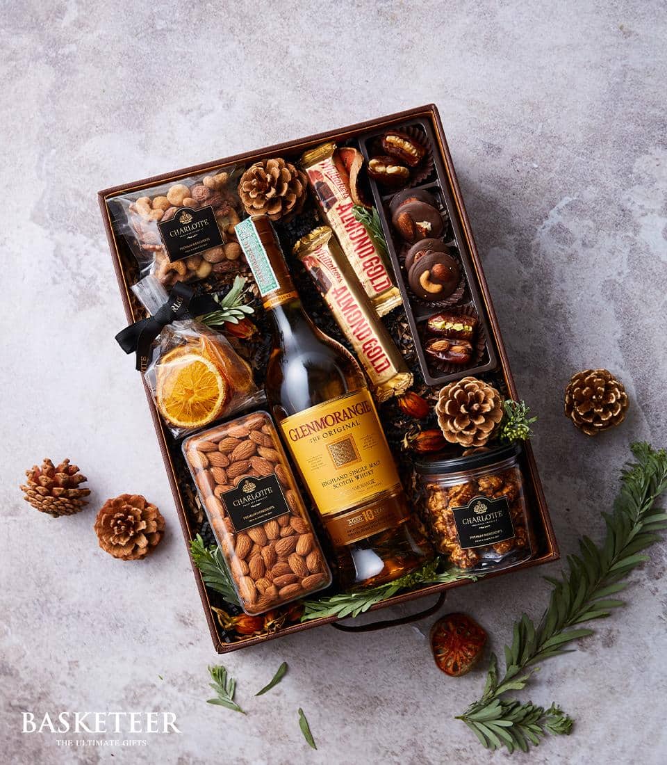 A luxurious gift basket curated by Basketeer features a refined bottle of Glenmorangie whiskey, alongside indulgent Chuaqette caramelized nuts. The assortment is completed with decadent Ferrero Rocher chocolates, dried orange slices, and assorted nuts encapsulated in elegant jars. Adorned with pine cones and lush evergreen sprigs, the basket exudes a celebratory charm and rests gracefully on a light grey surface. A tasteful ribbon displaying the Basketeer logo adds an exquisite finishing touch to this opulent presentation.