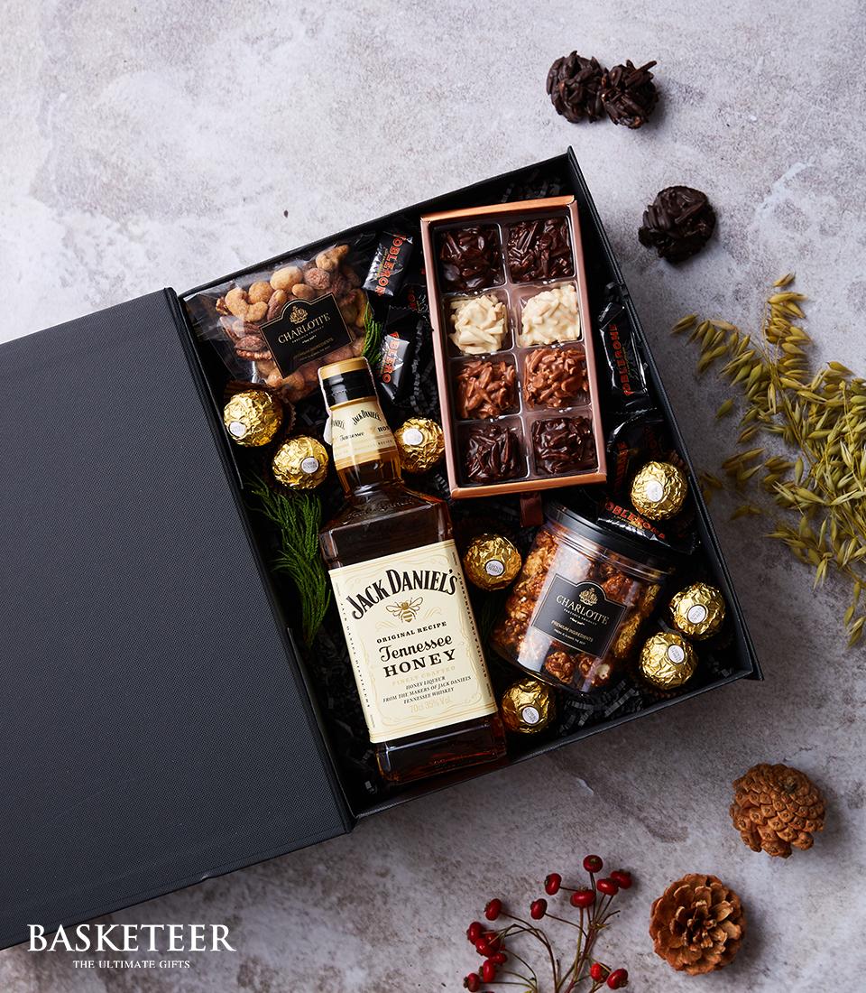 An elegant open gift box filled with luxurious indulgences, featuring a refined bottle of Jack Daniel's Tennessee Honey. Nestled beside are assorted premium chocolates, two chic jars brimming with delectable chocolate treats, and an array of divine Ferrero Rocher chocolates. Complementing these delights are an assortment of gourmet nuts. Each item is meticulously arranged within a sophisticated black box adorned with the prestigious Basketeer logo at the bottom left corner, making this a perfect blend of opulence and warmth.
