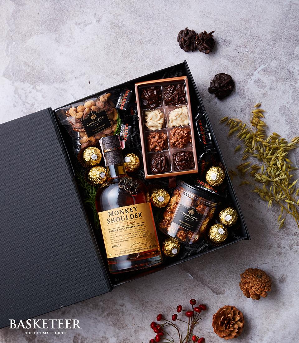 A sumptuous gift box featuring an exquisite bottle of Monkey Shoulder whiskey, elegantly paired with a selection of assorted chocolates nestled within a sophisticated tray. Delight in the indulgence of a jar filled with chocolate-covered nuts, accompanied by individual packages of Ferrero Rocher chocolates and an assortment of premium nuts. The scene is adorned with decorative pinecones, berries, and lush leaves for an added touch of festive opulence. The box proudly showcases the refined 