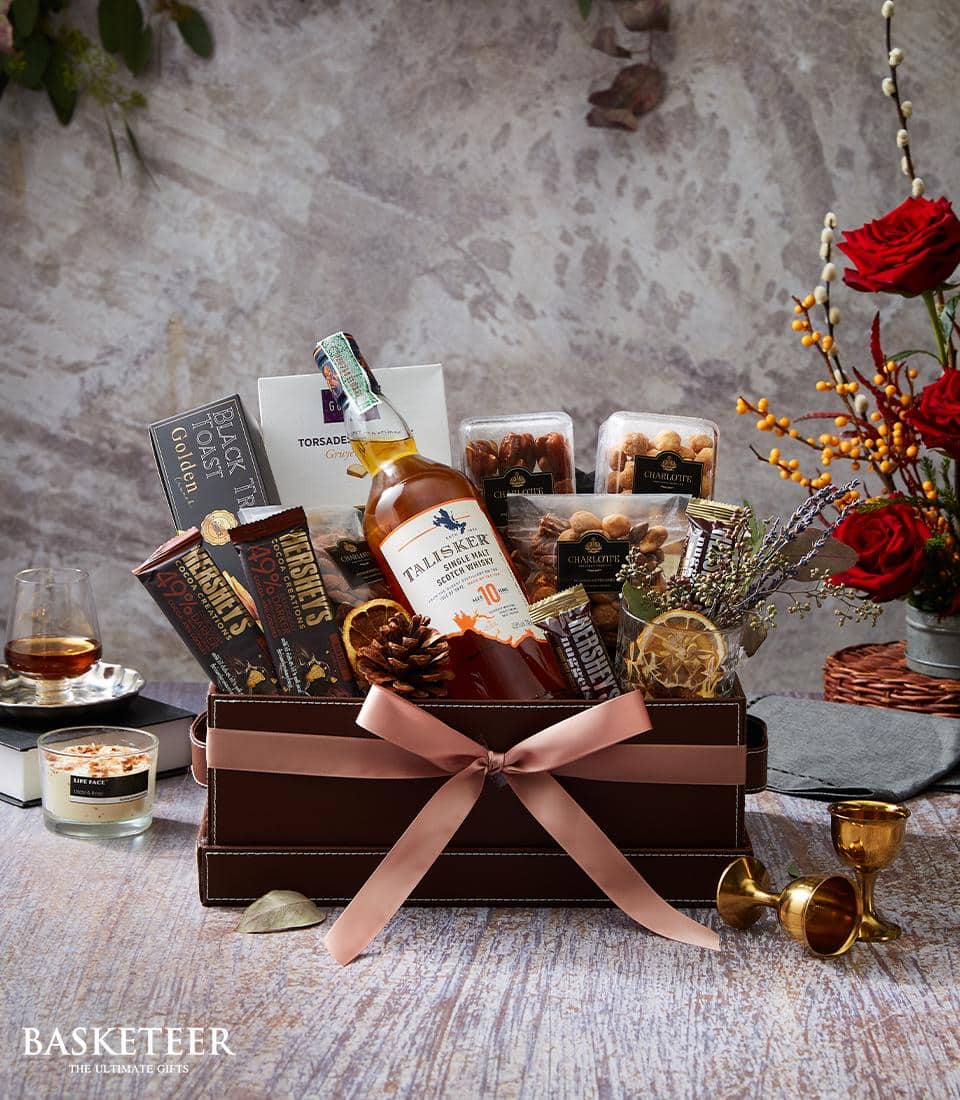 An exquisitely curated Basketeer gift basket presents a bottle of Trailseeker whiskey, surrounded by decadent gourmet chocolates, premium nuts, and an elegant bouquet of vibrant flowers. The ensemble is meticulously wrapped with a luxurious pink ribbon adorned with the Basketeer logo and paired with a softly glowing candle and two sophisticated glasses of whiskey—inviting indulgence and celebration in the epitome of luxury.