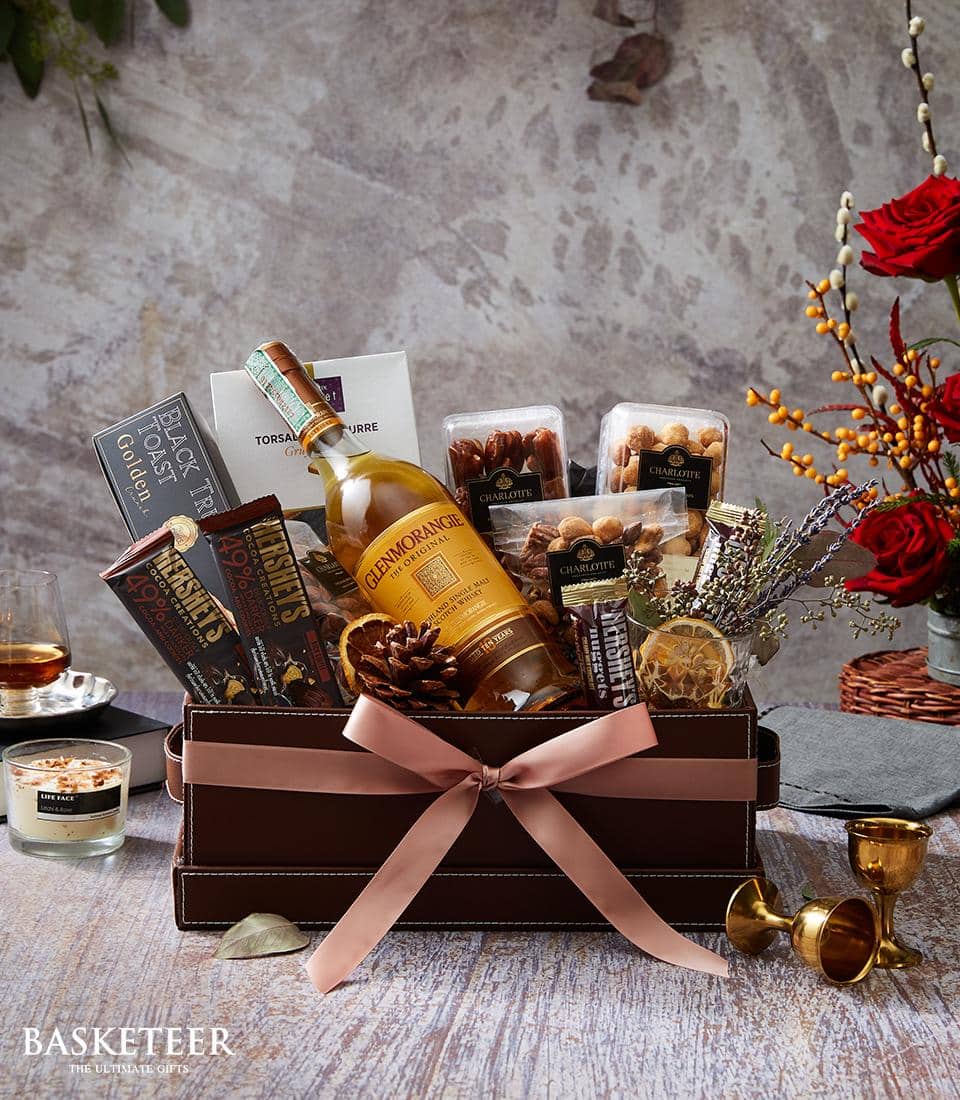 An opulent gift hamper by Basketeer, elegantly adorned with a pink ribbon bearing the Basketeer logo, brimming with a bottle of Glenmorangie whiskey, an assortment of fine chocolates, premium nuts, and gourmet snacks. The lavish basket is nestled on a refined wooden surface surrounded by a sophisticated glass of whiskey, flickering candlelight, gleaming gold goblets, and delicate flowers in the background.