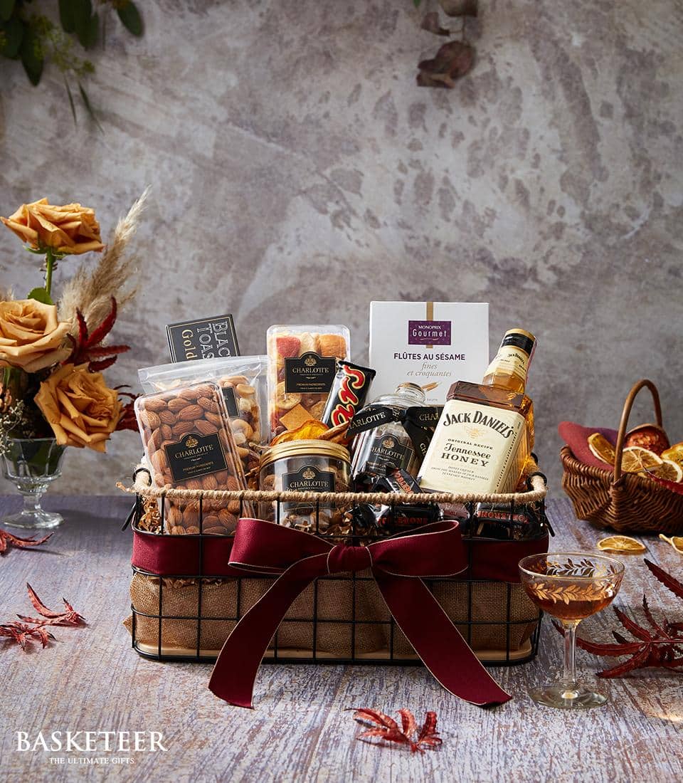 An exquisite gift basket adorned with a luxurious plaid cloth liner and an elegant red ribbon bow features premium Jack Daniel's whiskey, gourmet mixed nuts, delectable biscuits, savory pretzels, and indulgent chocolates. The Basketeer logo is prominently displayed on a sophisticated ribbon that graces the basket. In the foreground, a crystal glass of whiskey complements artfully arranged dried orange slices, while beautifully dried flowers in the background create a refined and inviting ambiance.