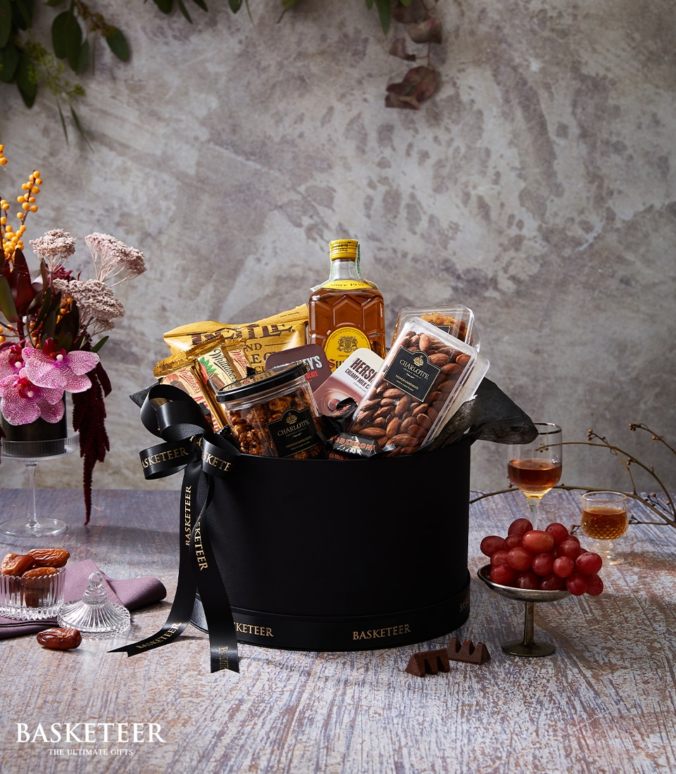 An opulent black gift basket by Basketeer, elegantly brimming with an assortment of premium snacks like rich chocolates, roasted almonds, delectable cookies, and a distinguished bottle of whiskey. This luxurious ensemble is finished with a signature Basketeer black ribbon. Complementing the lavish presentation are various vibrant flowers, a glass of fine whiskey, and a platter of succulent grapes arranged nearby.