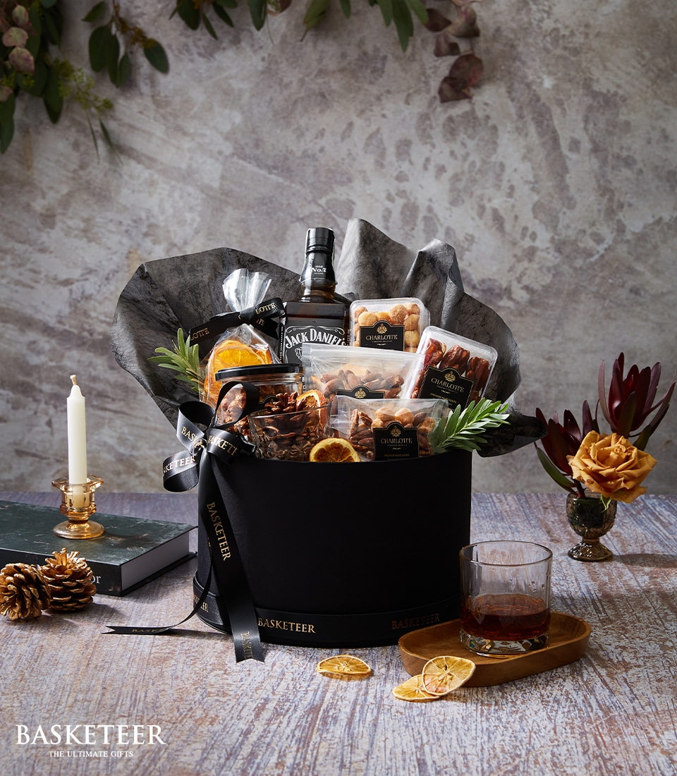 A sumptuous Basketeer gift basket overflowing with gourmet indulgences: Jack Daniel's whiskey, an assortment of premium nuts, decadent chocolates, and exquisite artisan treats. The ensemble is elegantly adorned with a sleek black ribbon featuring the Basketeer logo and thoughtfully positioned on a refined table alongside a glowing candle, a chic succulent, and a crystal glass of whiskey.