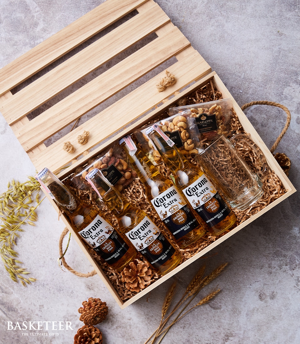 An exquisite wooden gift hamper with a charming slatted lid reveals three bottles of premium Corona Extra beer, two elegant sachets of assorted gourmet nuts, and a crystal-clear glass, all nestled amid artful shredded paper. The arrangement is accentuated by the natural beauty of dried wheat and rustic pine cones. A sophisticated touch includes the Basketeer logo adorning the ribbon and elegantly branded box. Text at the bottom left reads 