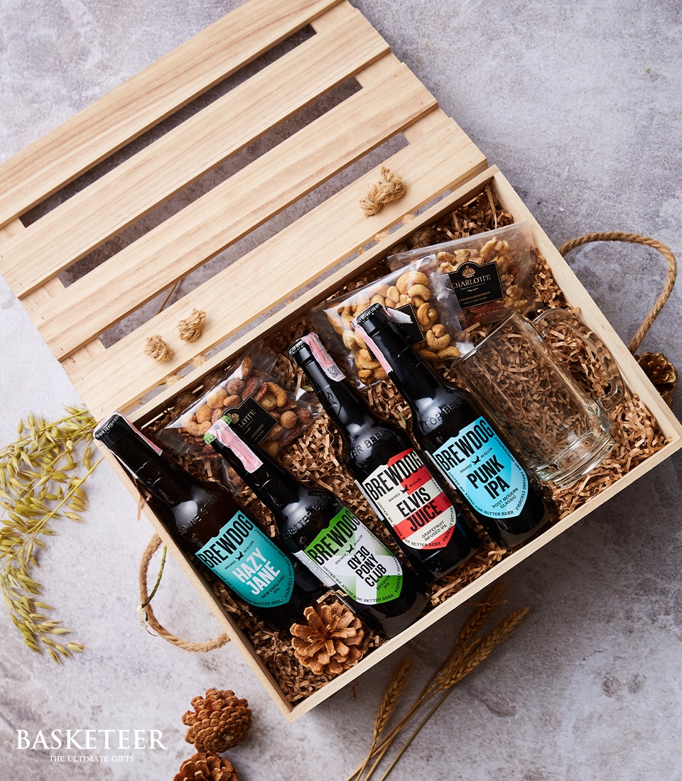 A luxurious wooden gift box from Basketeer, elegantly presented with four exquisite bottles of BrewDog beer, two gourmet packets of mixed nuts, and a pristine glass ready for use. The interior is meticulously lined with natural straw, artfully arranging each item to create an inviting display. This opulent gift set lies open on a textured surface, showcasing the premium Basketeer brand ribbon and logo that epitomize sophistication and indulgence.