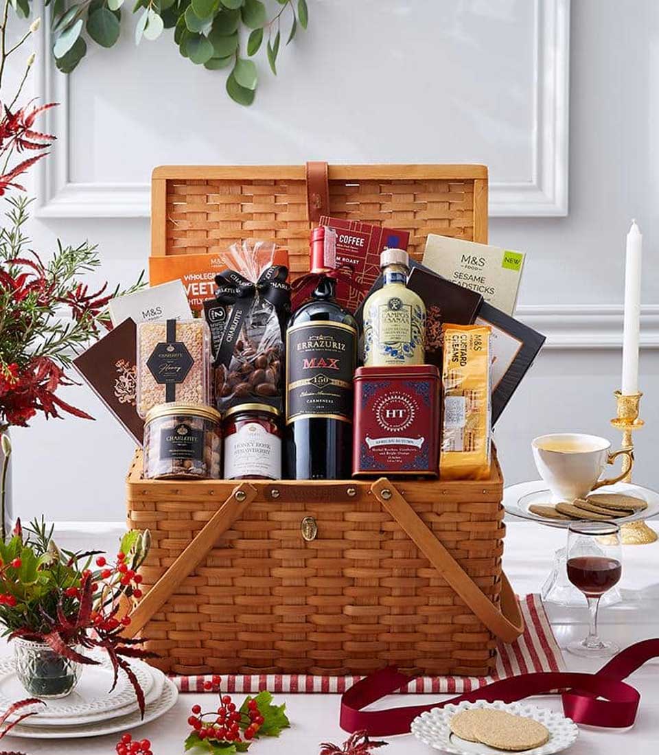 A luxurious Basketeer gift basket brimming with gourmet delights takes center stage, featuring a finely selected bottle of wine, artisanal chocolates, delectable cookies, premium tea, assorted nuts, and exquisite preserves. Nestled on an elegantly adorned festive table with a glowing candle and a sophisticated teacup beside it. The basket is accentuated by a chic red and white ribbon bearing the Basketeer logo. Enveloped in holiday charm, lush greenery and vibrant red berries frame this inviting scene.