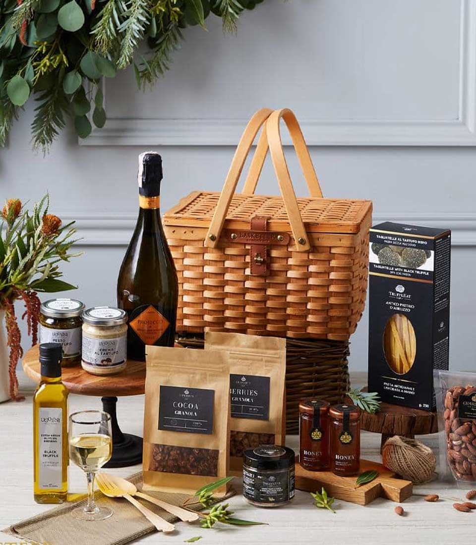 A luxurious wicker gift basket from Basketeer elegantly rests on a polished table, brimming with an assortment of gourmet delights. The sophisticated collection includes a bottle of exquisite sparkling wine, a lavish jar of golden honey, artisan pasta, premium olive oil, rich cocoa granola, and an array of fragrant spices. A gracefully filled glass of white wine adds to the opulent arrangement. Lush greenery provides an enchanting backdrop, enhancing the indulgent ambiance. A delicate ribbon adorned with the distinguished Basketeer logo completes this sumptuous scene.
