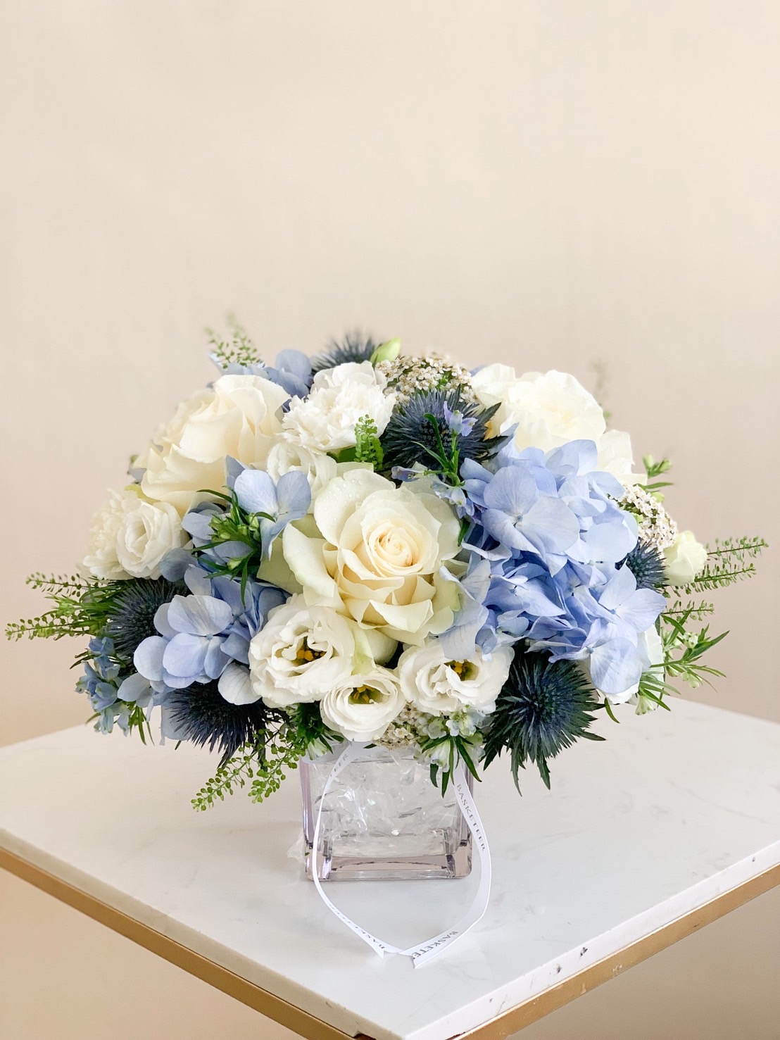 Elegant vase arrangement with white roses, blue hydrangeas, and greenery in a square glass vase.