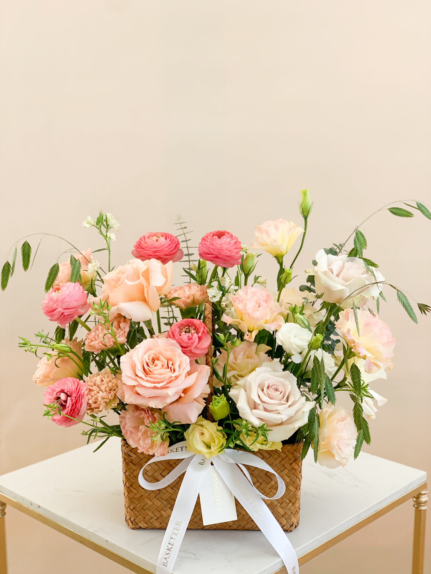 Woven basket filled with pastel-colored roses, ranunculus, and carnations, adorned with a white Basketeer ribbon.