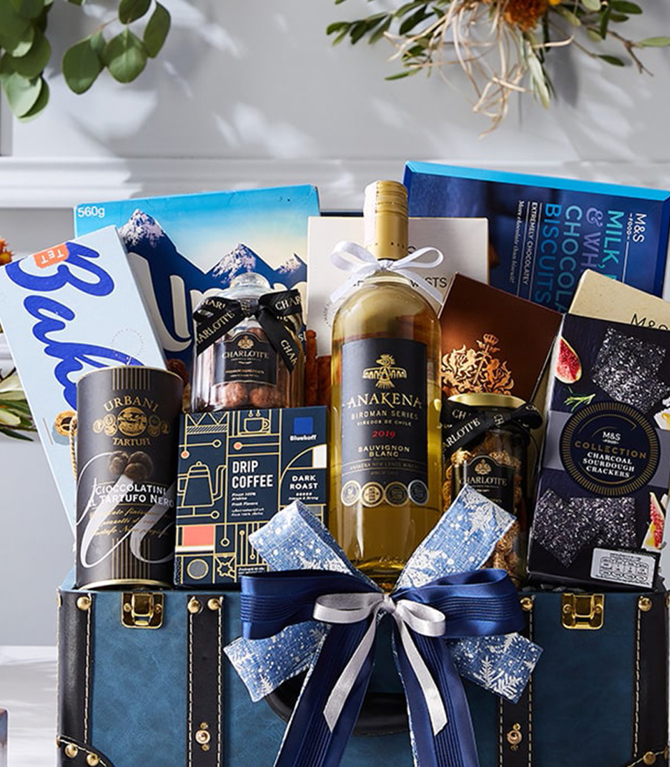 A luxurious blue-themed gourmet gift hamper featuring white wine, fine chocolates, premium coffee, and indulgent snacks, elegantly presented in a vintage-style leather trunk with a festive bow.