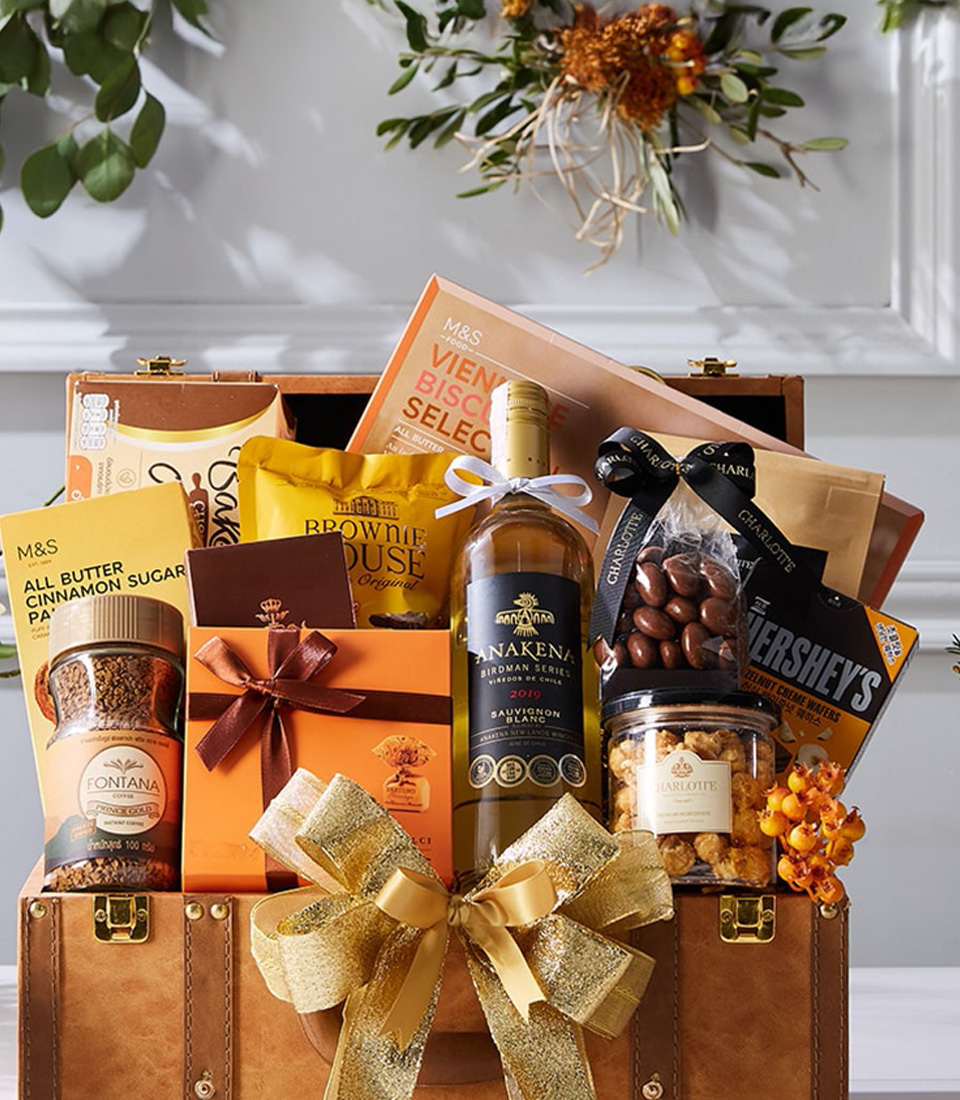 An opulent gift hamper by Basketeer, elegantly brimming with gourmet treasures like fine wine, artisanal chocolates, premium nuts, and delectable snacks. These exquisite treats are meticulously arranged within a vintage-style trunk adorned with a lavish gold ribbon that proudly showcases the Basketeer logo. Encircled by charming autumn-themed decor on a polished wooden table, this scene exudes warmth and luxury. At the bottom of this radiant display reads 