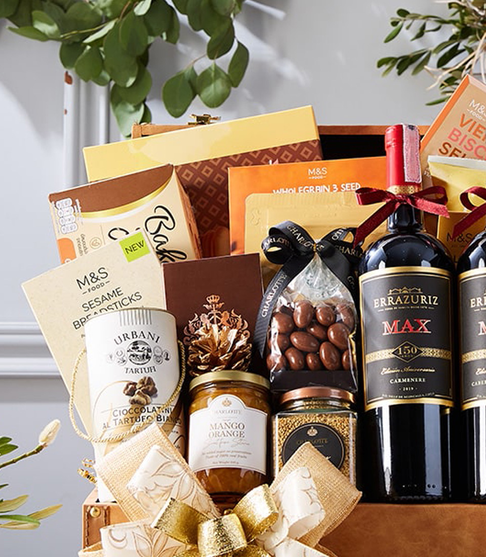 An opulent gift basket featuring two exquisite bottles of red wine, an array of decadent chocolates, premium cookies, artisanal crackers, and other gourmet delights. All these treasures are exquisitely arranged in a luxurious brown leather case adorned with a lavish gold bow. The scene is completed with a softly glowing candle and festive decorations set on a pristine white table. The epitome of indulgence from Basketeer, marked by their recognizable ribbon and crafted to impress.