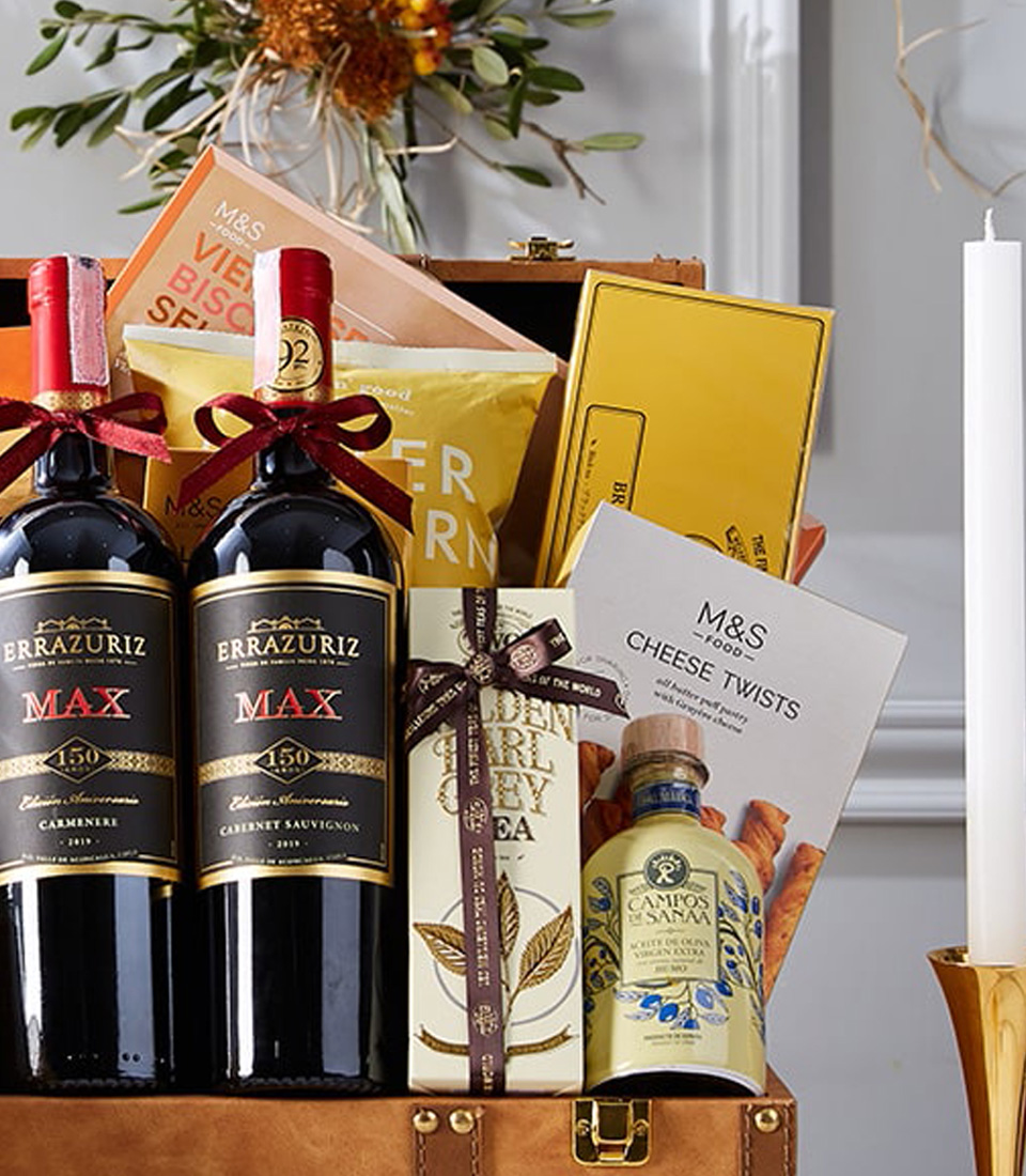 An opulent gift basket featuring two exquisite bottles of red wine, an array of decadent chocolates, premium cookies, artisanal crackers, and other gourmet delights. All these treasures are exquisitely arranged in a luxurious brown leather case adorned with a lavish gold bow. The scene is completed with a softly glowing candle and festive decorations set on a pristine white table. The epitome of indulgence from Basketeer, marked by their recognizable ribbon and crafted to impress.