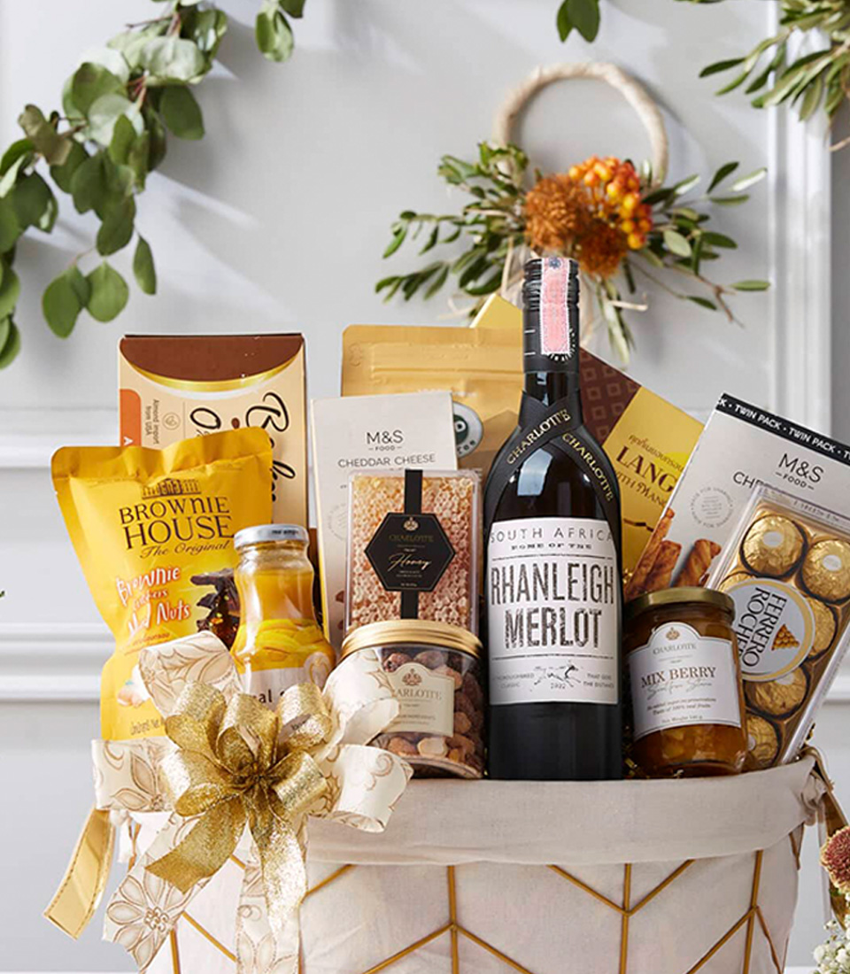 A gift basket filled with various gourmet items including a bottle of red wine, chocolate truffles, mustard, olives, savory biscuits, and sauces. The basket is adorned with a ribbon and placed on a white table with festive greenery and flowers in the background.