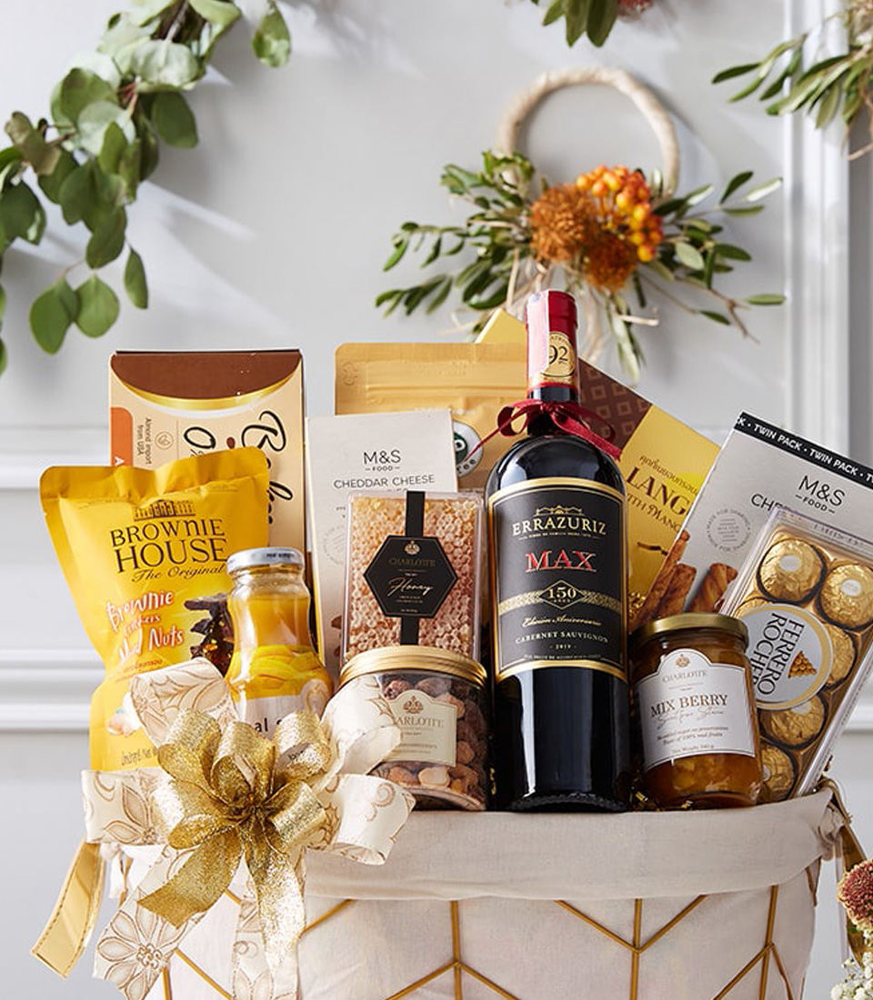 A sumptuous Basketeer Vino Snack Affair Basket overflowing with fine wine, decadent chocolates, and a curated selection of gourmet treats, elegantly tied with a shimmering gold ribbon and festive embellishments. This luxurious ensemble is artfully presented on a pristine white surface, complemented by an exquisite wine glass and delicate floral arrangements in petite vases. The distinguished 