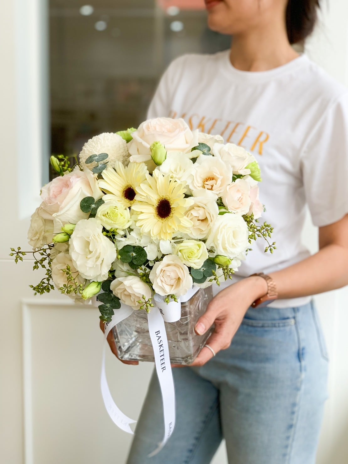 A person wearing a white T-shirt and blue jeans holds a glass vase filled with a bouquet of white and pastel yellow flowers, including roses and daisies. The vase has a white ribbon with text on it, and the person is facing slightly to the right.