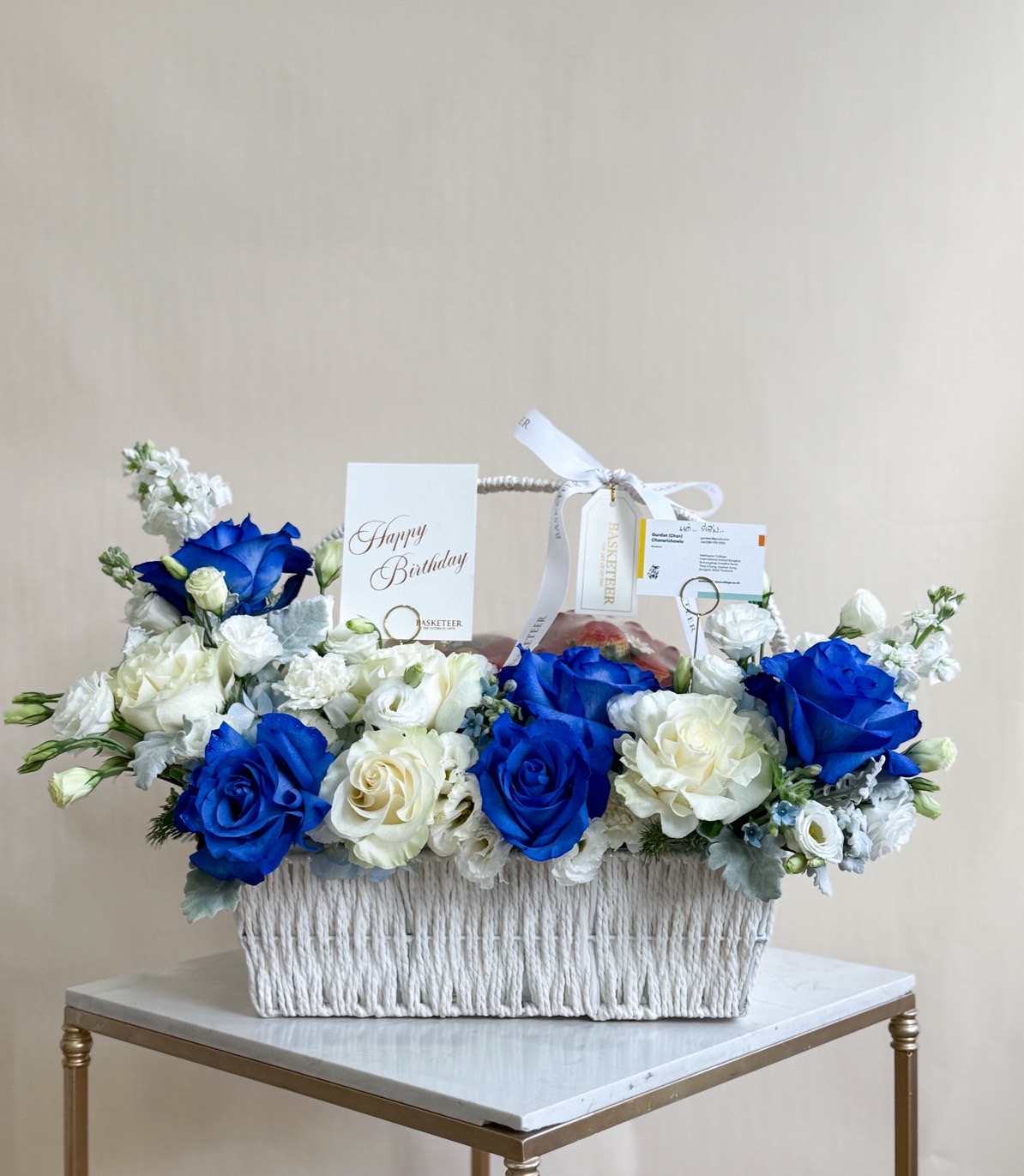 Elegant white basket arrangement with blue roses, white roses, carnations, lisianthus, and foliage, decorated with Basketeer ribbon and card.