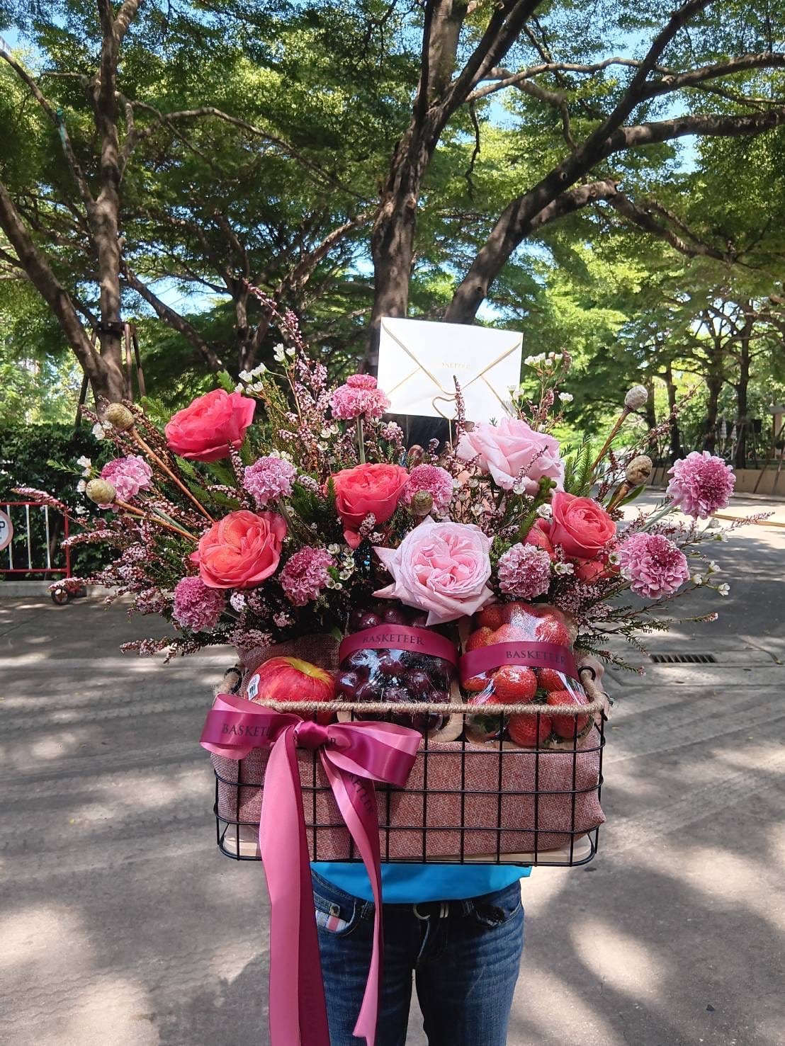 A person in a blue shirt is holding a Sweetheart Fruit Floral Basket filled with pink and red flowers, apples, and adorned with pink ribbons. The background features trees and a sunny outdoor setting. A white envelope is placed among the flowers.