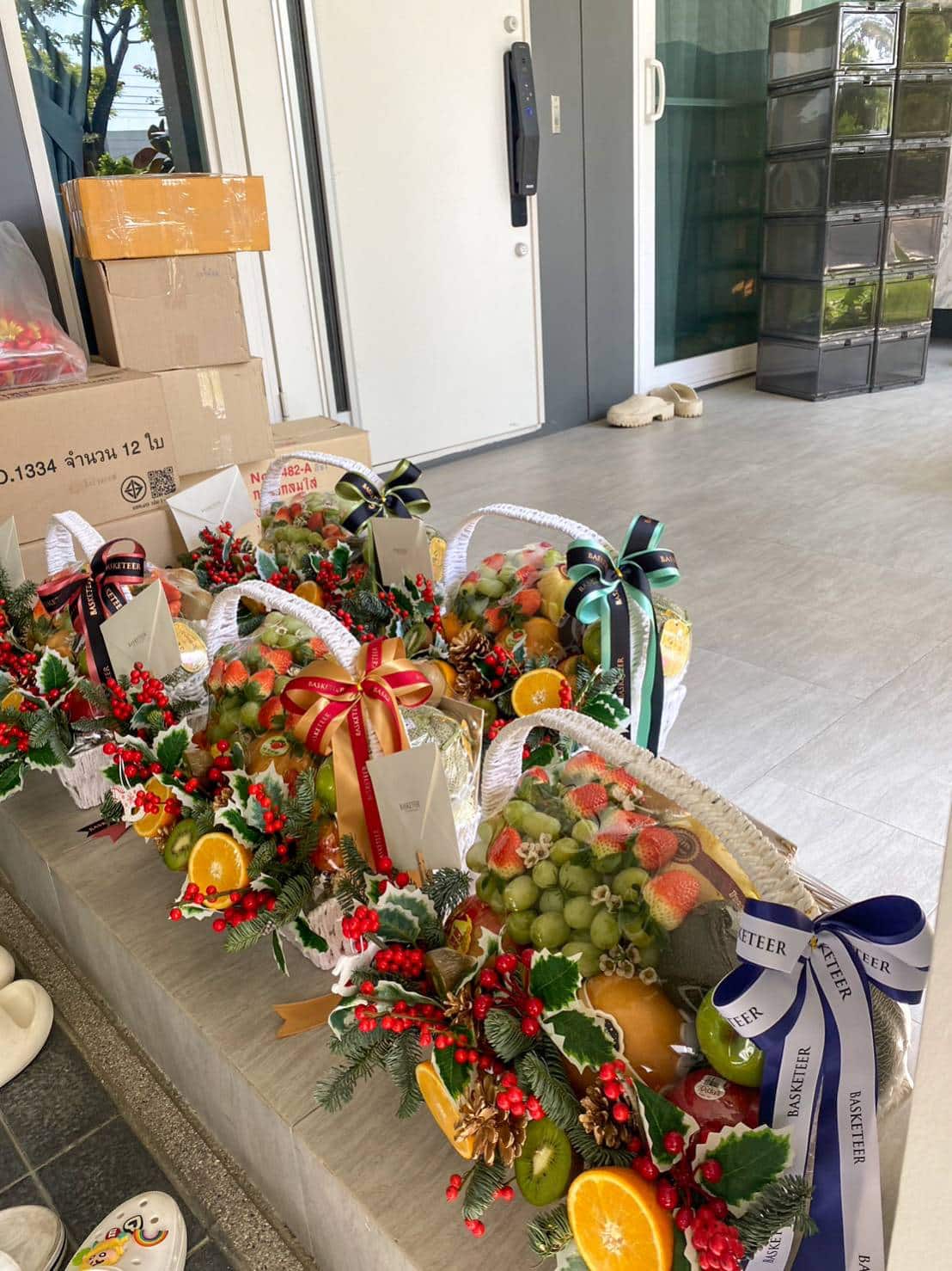 Four festive Premium Fruits Greetings Baskets filled with assorted fruits, adorned with colorful ribbons and Christmas-themed decorations, are displayed on a porch. A stack of cardboard boxes and a closed door are in the background, and a dog is lying down in the distance.